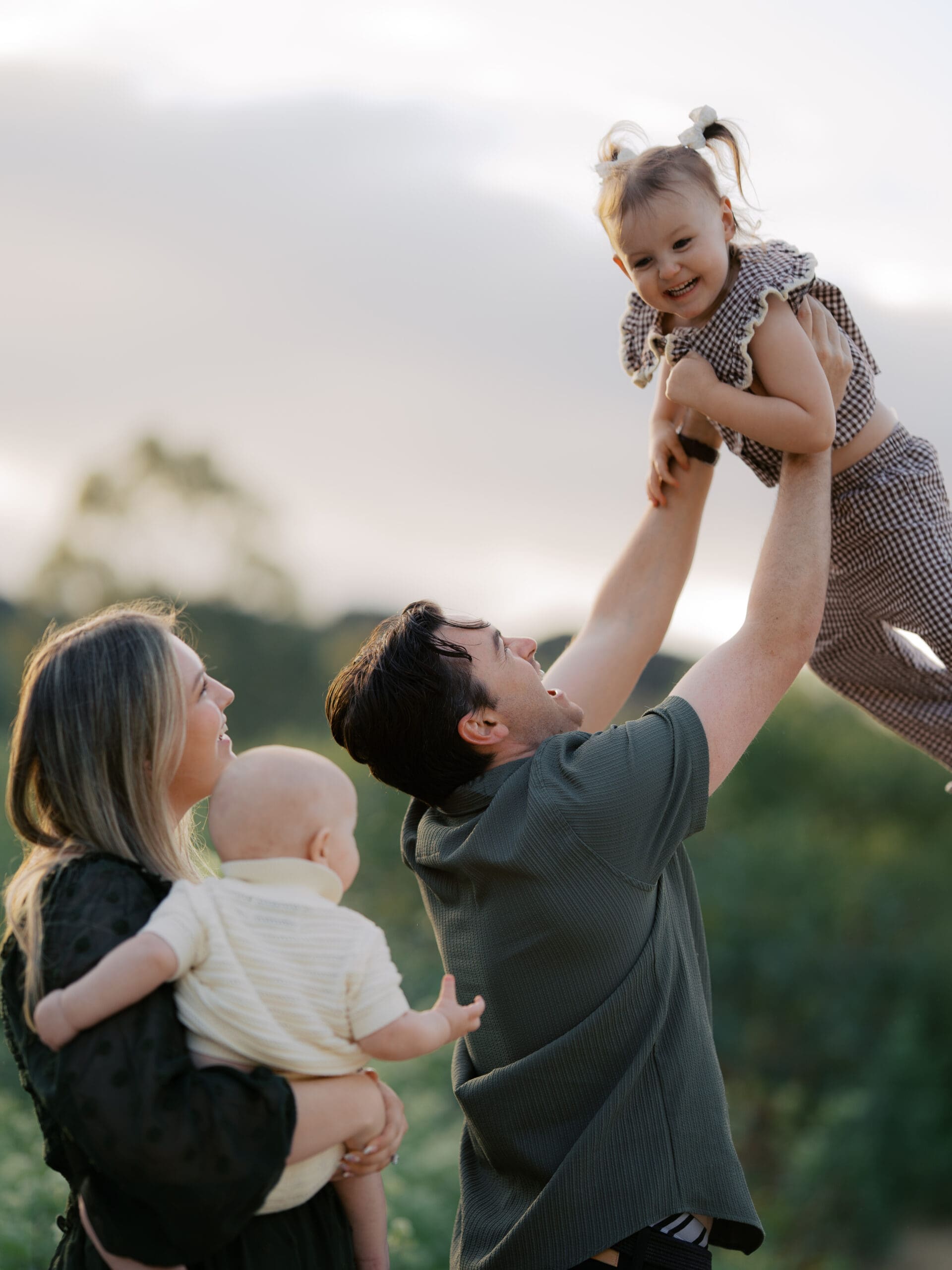 A mother holiding a baby watching the father swing their happy toddler up in the air during golden hour photoshoot by camarillo family photographer.