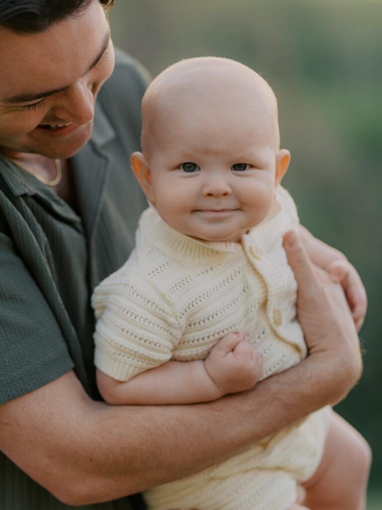 Smiling baby in cream knit romper being held by father during Camarillo family photography session in natural outdoor setting