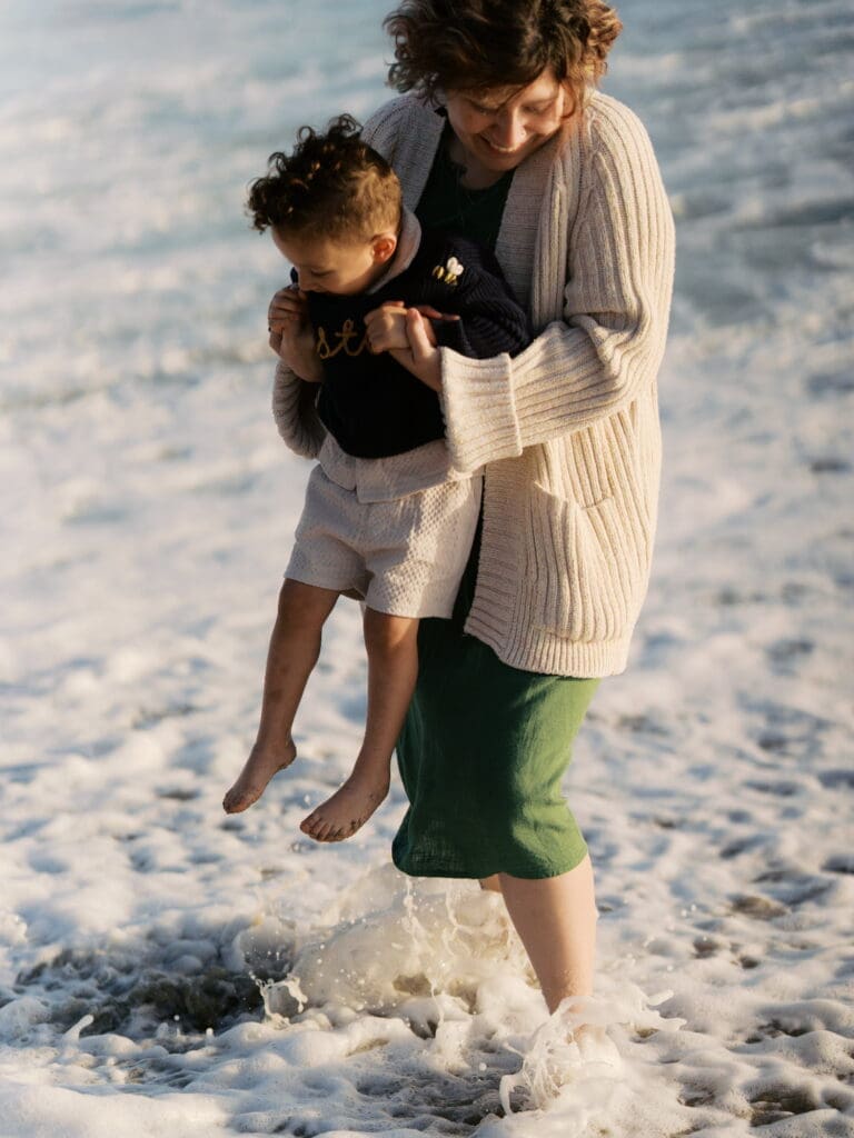 A mother in a green dress playfully lifts her child up out of the crashing surf.