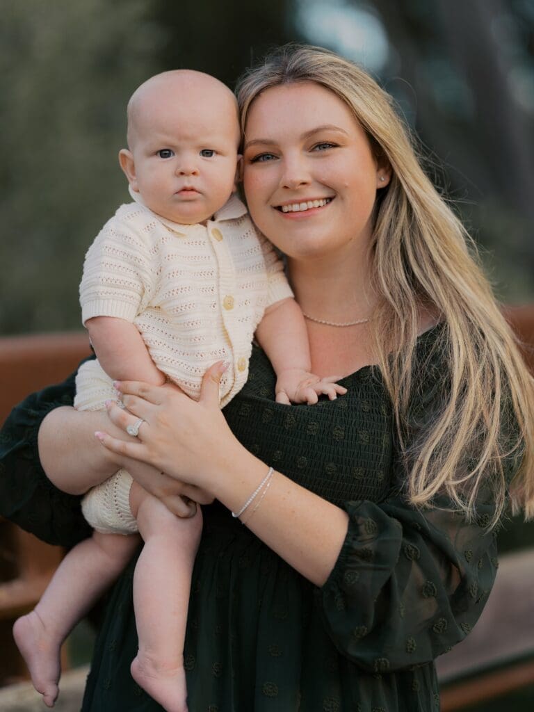 Smiling mother in dark green dress holding baby in cream knit romper during outdoor family photography session in Camarillo