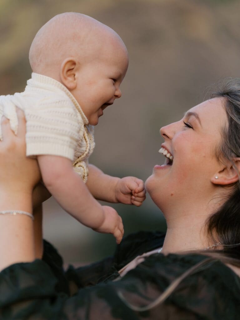 Mother lifting laughing baby in cream knit romper face-to-face during golden hour family session in Ventura County