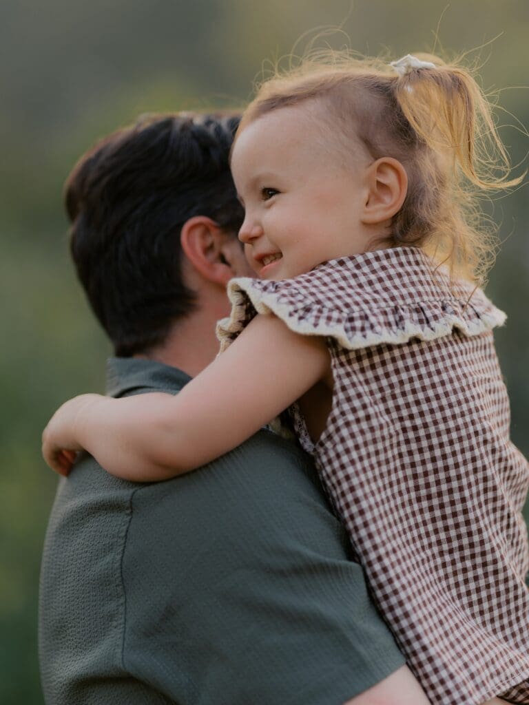 Smiling toddler girl in gingham ruffle romper hugging father's shoulder during Hill Canyon Trail family photography session in Camarillo