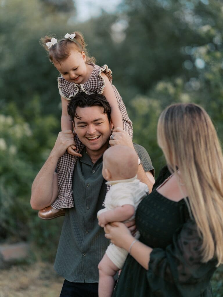 Happy family of four laughing outdoors with toddler on father's shoulders and mother holding baby during Hill Canyon Trail family photography session
