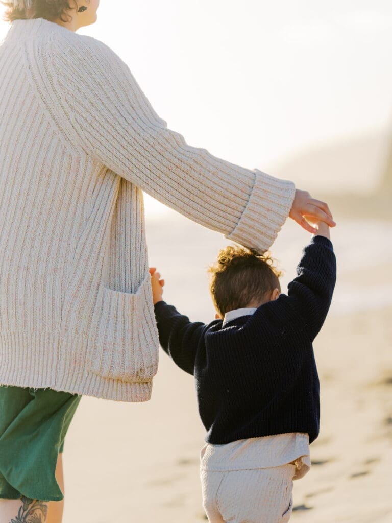 A young boy leads holds his mothers hands up over his head while leading her down the beach.