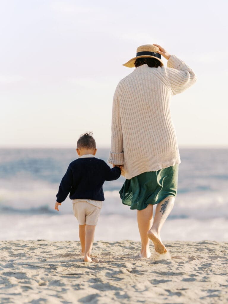 A woman wearing a cream knit cardigan over a green dress and a straw boater hat with a navy ribbon stands on a beach holding a curly-haired toddler dressed in a navy embroidered sweater. They are walking together on the beach towards the water while holding hands. The mother holds her hat on her head with the other hand.