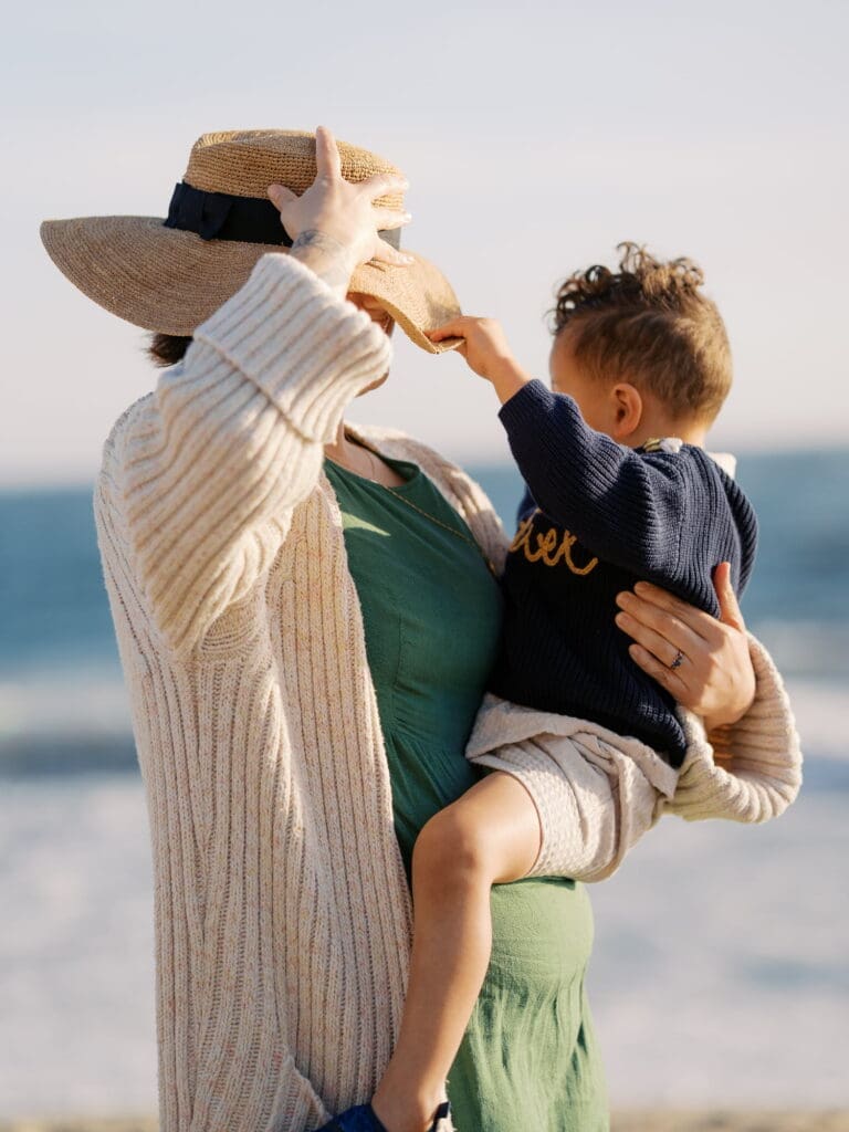 A woman wearing a cream knit cardigan over a green dress and a straw boater hat with a navy ribbon stands on a beach holding a curly-haired toddler dressed in a navy embroidered sweater. The child reaches up to grab the brim of her hat. The ocean is softly blurred in the background under a pale sky, suggesting late-afternoon light.