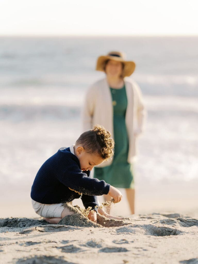 A woman wearing a cream knit cardigan over a green dress and a straw boater hat with a navy ribbon stands out of focus in the background while watching her child play in the sand. The child is wearing a navy sweater and is the focal point of the photo.