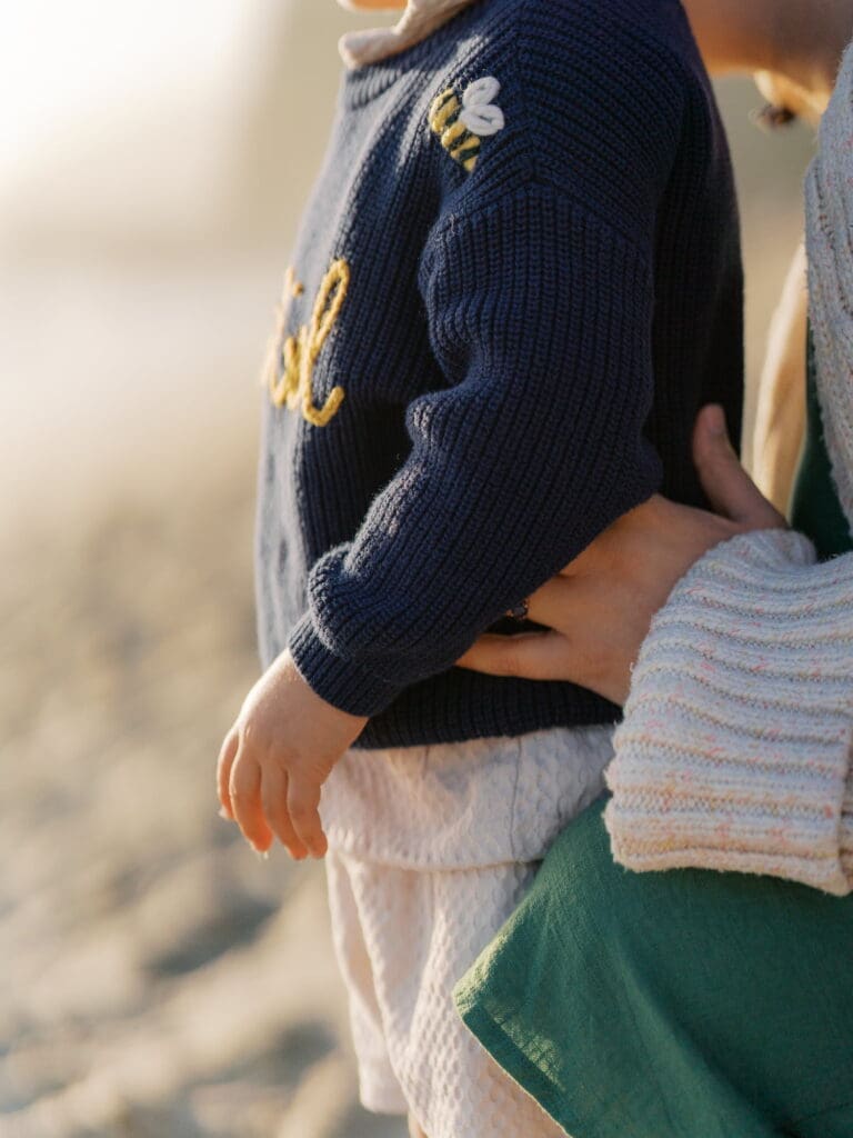 A close up image of a mother at the beach holding the waist of her child while looking out at the sunset over the ocean.