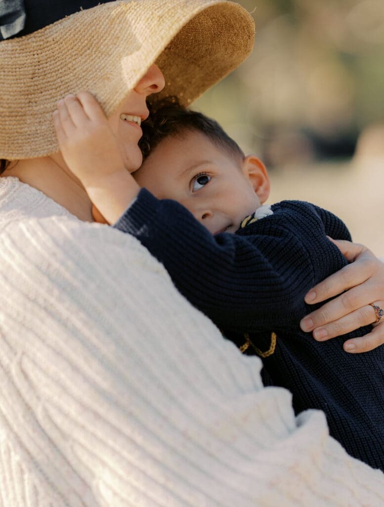 A mother wearing a straw hat cuddles her child on her chest. The child is reaching up and holding his mothers hat while looking up to the sky.