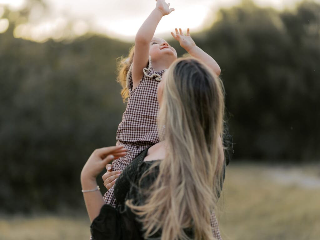 Mother lifting toddler girl in gingham romper above her head during golden hour family photo session in Ventura County