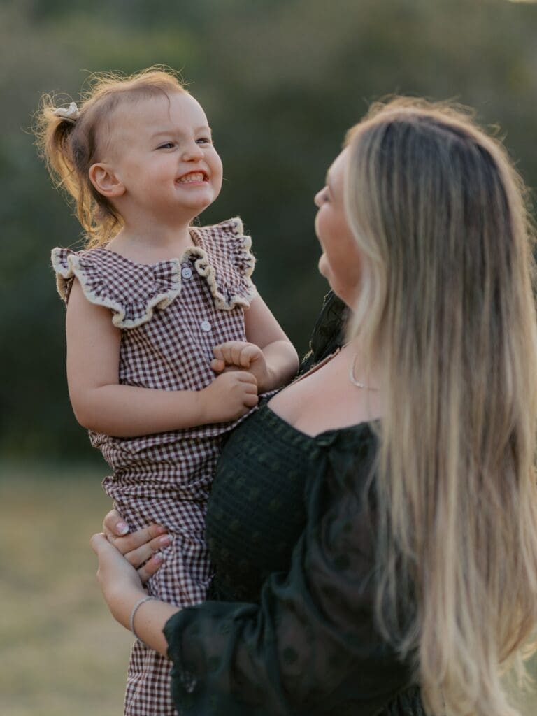 Laughing toddler girl in gingham ruffle romper held by mother during Ventura County outdoor family photography session at sunset
