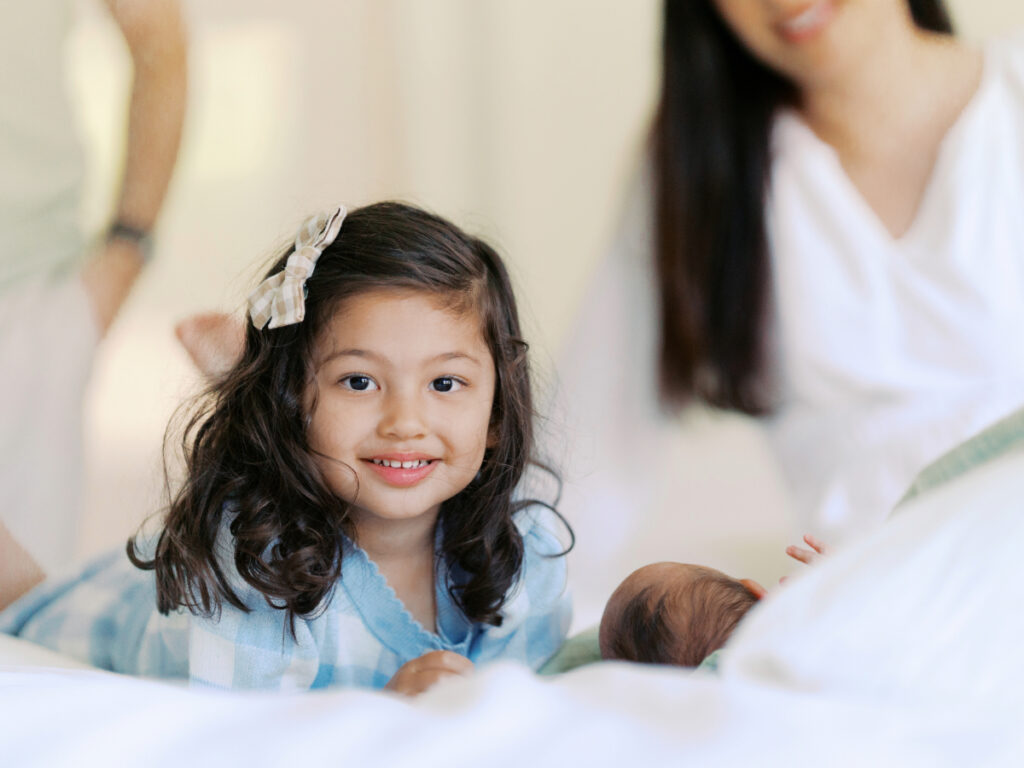 Little girl with dark curly hair looks at the camera while laying on a bed next to a newborn baby boy.