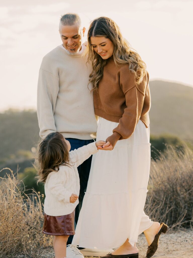 Mom and dad smiling down at their toddler holding mom’s hand on a hilltop trail in Thousand Oaks at golden hour